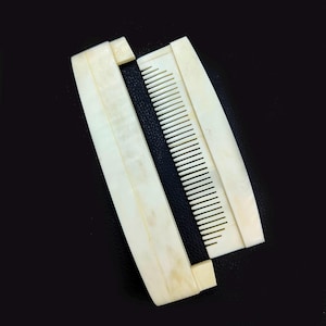 Puede incluir: Peine de color marfil con funda protectora, abierto sobre una superficie negra. Los dientes del peine son visibles y la funda es rectangular con bordes redondeados. El peine y la funda parecen estar hechos de un material sólido.