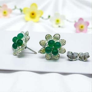 May include: A pair of silver-coloured flower-shaped stud earrings with green gemstone accents. The earrings have a cluster of green stones at the centre and are displayed with earring backs. The background features decorative flowers.