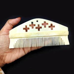 Puede incluir: Peine de color marfil con una parte superior decorativa con recortes en forma de cruz. El peine tiene dientes muy juntos y se sostiene en una mano sobre un fondo oscuro. El peine probablemente se usa para el cuidado del cabello.