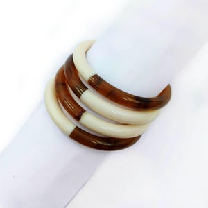 Può includere: Quattro braccialetti impilati. Ogni braccialetto è una combinazione di crema e marrone. La sezione marrone ha un motivo a scaglie di tartaruga. I braccialetti sono su sfondo bianco. Gioielli.