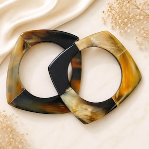 Peut inclure: Deux bracelets carrés avec une finition lisse et polie, présentant un motif marbré dans des tons de noir, ambre et crème. Les bracelets ont des ouvertures intérieures arrondies et des bords extérieurs angulaires, offrant un aspect naturel.