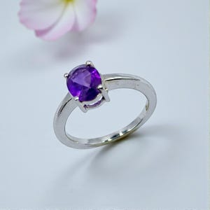 May include: A silver ring featuring a large, oval-cut, deep purple amethyst gemstone. The gemstone is held in place by four prongs. The ring band is a simple, polished silver. A pink flower is in the background.