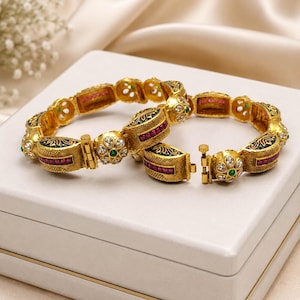 May include: Two gold-toned hinged bangles featuring intricate textured metalwork, inlaid with rectangular red stones, clear white crystals, and small green accents. Each bracelet includes a secure pin-style clasp mechanism and a floral-inspired design.