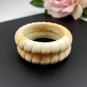 Puede incluir: Dos brazaletes de color marfil con un diseño acanalado. Los brazaletes están apilados, mostrando sus detalles tallados. El fondo es una superficie oscura con una rosa rosa borrosa. Cada brazalete tiene un diámetro de aproximadamente 7 cm.