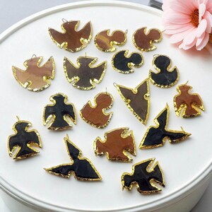 May include: A selection of pendant charms in bird and abstract shapes. The charms are in shades of brown and black, with gold-coloured edging. They are displayed on a white surface, with a pink flower in the background.