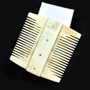 Könnte beinhalten: Ein cremefarbener, doppelseitiger Kamm mit feinen Zähnen. Der Kamm besteht aus einem hellen Material, möglicherweise Knochen oder Elfenbein, und wird mit kleinen Nieten zusammengehalten. Der Hintergrund ist schwarz.