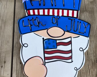 May include: A wooden door hanger shaped like a gnome with a blue hat that says "Happy 4th of July". The gnome is holding a small American flag in its hand.