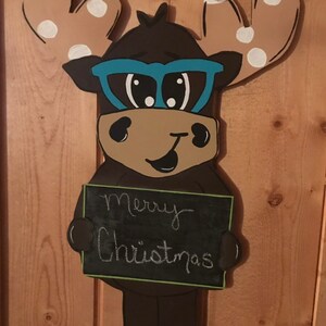 May include: A brown moose with blue glasses and brown antlers with white polka dots holds a chalkboard sign that says "Merry Christmas".