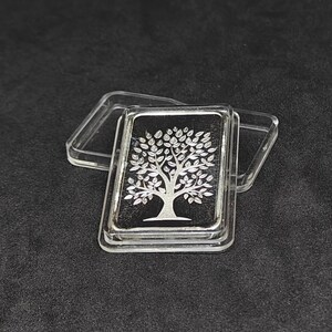 May include: A silver bar with a tree design on a black background, encased in a clear plastic case. The tree is detailed with leaves and branches. The bar is rectangular and has a shiny finish.