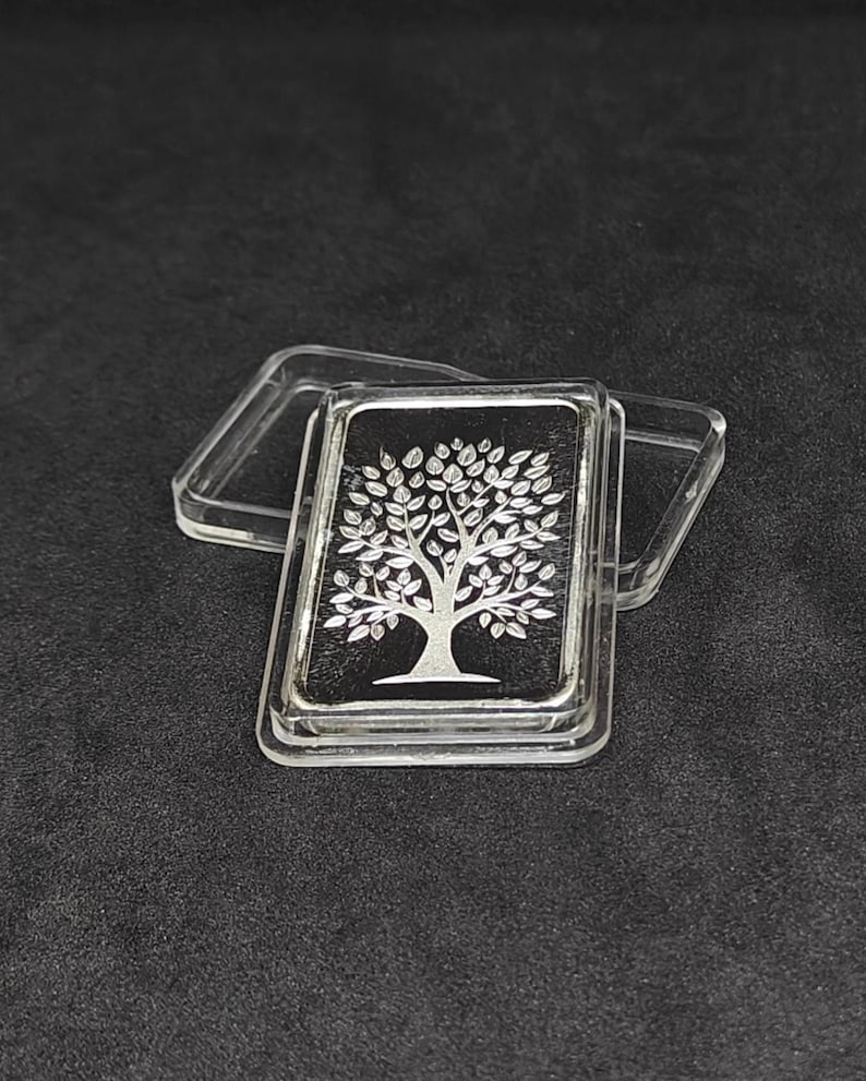 May include: A silver bar with a tree design on a black background, encased in a clear plastic case. The tree is detailed with leaves and branches. The bar is rectangular and has a shiny finish.