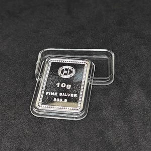 Puede incluir: Una barra rectangular de plata fina de 10 g, con una pureza de 999,9, se muestra en una caja de plástico transparente. La barra de plata tiene un logotipo y está sobre un fondo oscuro.