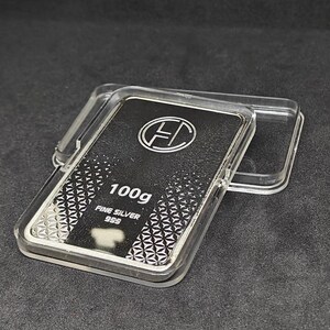 May include: A silver bar, weighing 100g, encased in a clear plastic container. The bar is marked with the text "FINE SILVER 999" and a logo. The bar has a geometric pattern on the bottom half.