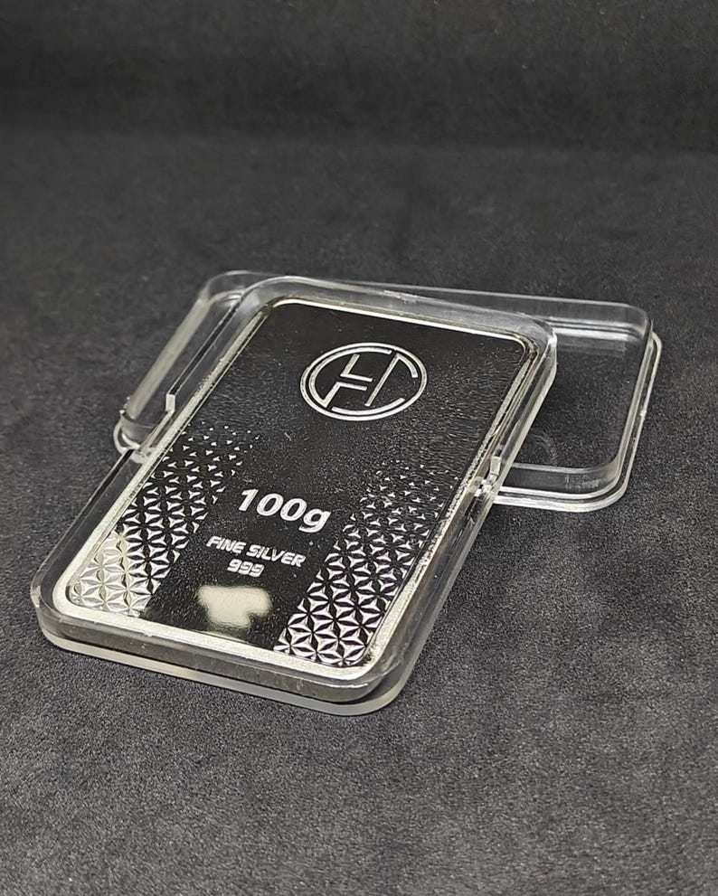 May include: A silver bar, weighing 100g, encased in a clear plastic container. The bar is marked with the text "FINE SILVER 999" and a logo. The bar has a geometric pattern on the bottom half.