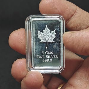 May include: A rectangular silver bar with a maple leaf design. The bar is marked "5 GMS FINE SILVER 999.9". The bar is held in a clear plastic case.