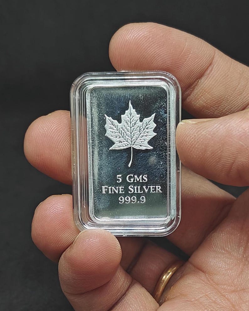 May include: A rectangular silver bar with a maple leaf design. The bar is marked "5 GMS FINE SILVER 999.9". The bar is held in a clear plastic case.