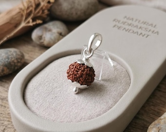 May include: A natural rudraksha pendant featuring a round, textured brown seed bead encased in polished silver caps with a silver bail. The pendant is displayed on a light beige oval stand with the text NATURAL RUDRAKSHA PENDANT printed on it.