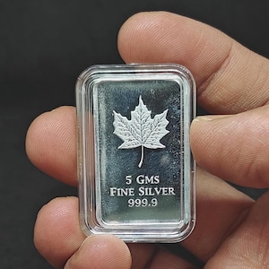 May include: A silver bar featuring a maple leaf design, encased in clear plastic. The bar is marked with "5 GMS FINE SILVER 999.9". The bar weighs 5 grams.