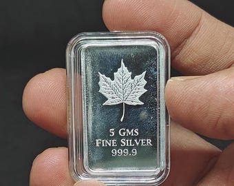 May include: A rectangular silver bar with a maple leaf design. The bar is encased in a clear plastic case. The text on the bar reads "5 GMS FINE SILVER 999.9".