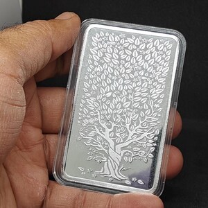 K&ouml;nnte beinhalten: Ein rechteckiger Silberbarren mit einem Baumdesign, umh&uuml;llt von einer transparenten Kunststoffh&uuml;lle. Der Baum weist detaillierte Bl&auml;tter und &Auml;ste auf. Der Barren wird in einer Hand vor dunklem Hintergrund gehalten.