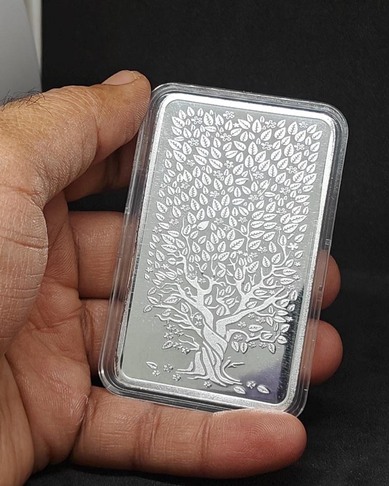 K&ouml;nnte beinhalten: Ein rechteckiger Silberbarren mit einem Baumdesign, umh&uuml;llt von einer transparenten Kunststoffh&uuml;lle. Der Baum weist detaillierte Bl&auml;tter und &Auml;ste auf. Der Barren wird in einer Hand vor dunklem Hintergrund gehalten.