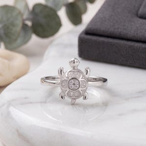May include: A silver-toned ring featuring a turtle-shaped centerpiece. The turtle shell is decorated with a floral pattern and small, clear, sparkling stones. The ring band is smooth and polished, showcasing a detailed design of the turtle's head, four legs, and a small tail extending from the central shell motif.