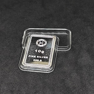 Puede incluir: Una barra rectangular de plata fina de 10g, marcada "FINE SILVER 999.9", se encuentra en una funda de plástico transparente. La barra tiene una superficie brillante y un borde decorativo. La funda está abierta, sobre un fondo oscuro.
