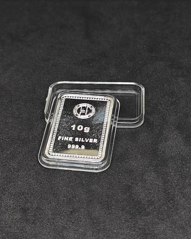 May include: A rectangular silver bar, marked "10g FINE SILVER 999.9", rests in a clear plastic case. The bar has a reflective surface and a decorative border. The case is open, revealing the bar against a dark background.