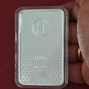 K&ouml;nnte beinhalten: Rechteckiger Silberbarren, 100g, in einer transparenten Kunststoffh&uuml;lle. Der Barren hat ein rundes Logo und den Text "FINE SILVER 999.9". Der Hintergrund ist rot.