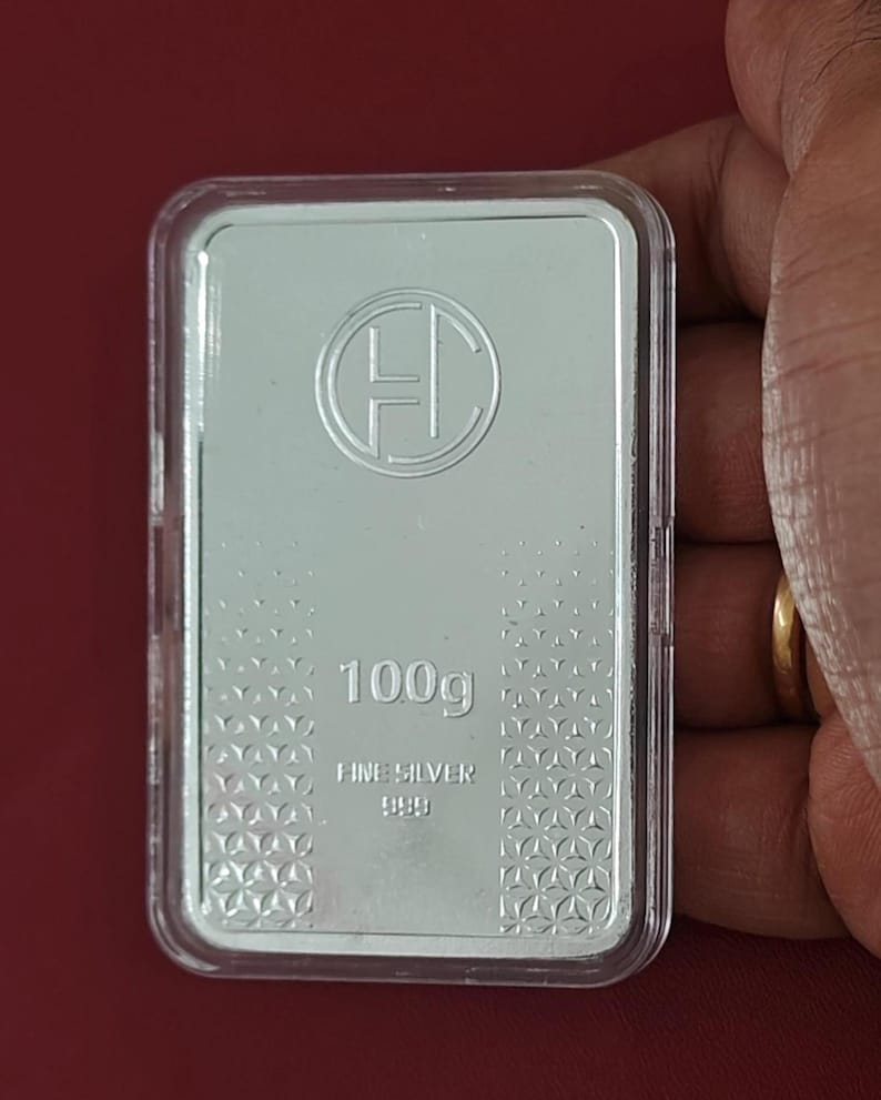 K&ouml;nnte beinhalten: Rechteckiger Silberbarren, 100g, in einer transparenten Kunststoffh&uuml;lle. Der Barren hat ein rundes Logo und den Text "FINE SILVER 999.9". Der Hintergrund ist rot.