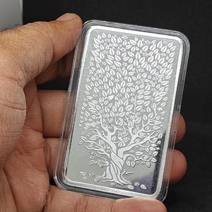 May include: A rectangular silver bar with a detailed tree of life design, held in a clear plastic case. The tree features a textured trunk, branches, and numerous leaves. The bar is held in a hand against a dark background.