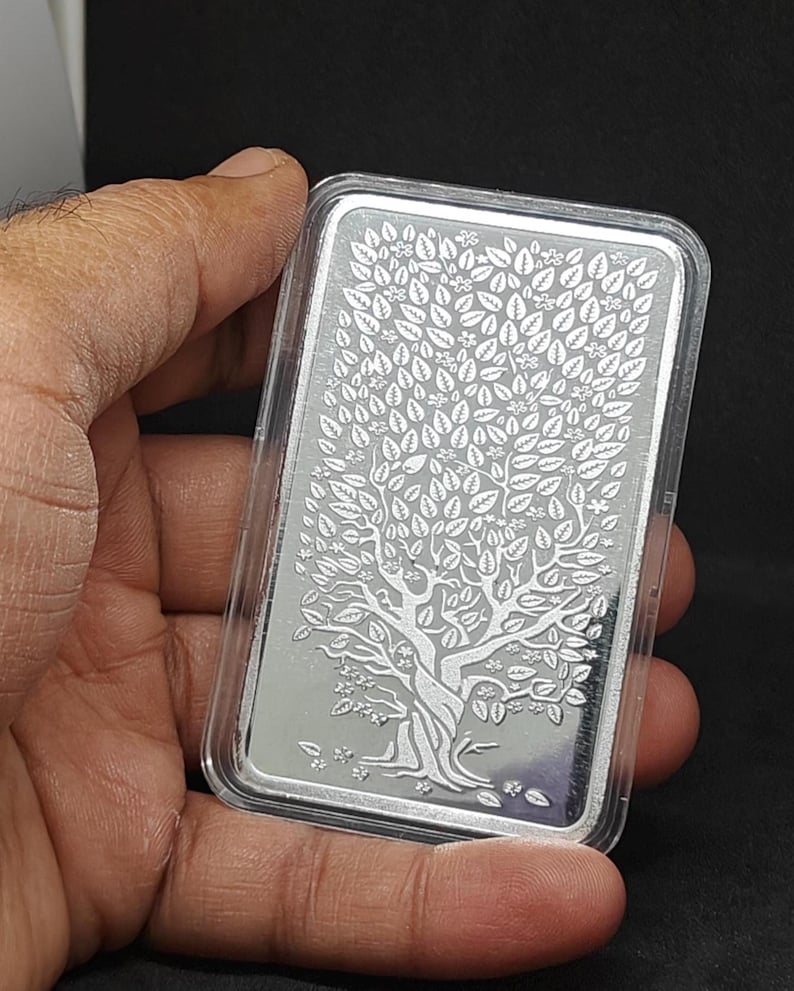 May include: A rectangular silver bar with a detailed tree of life design, held in a clear plastic case. The tree features a textured trunk, branches, and numerous leaves. The bar is held in a hand against a dark background.
