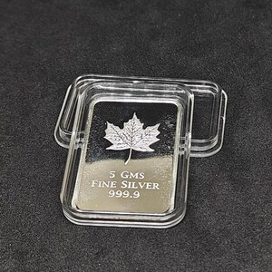 May include: A rectangular silver bar with a maple leaf design, encased in a clear plastic case. The bar is marked with "5 GMS FINE SILVER 999.9". The case is open, revealing the silver bar on a dark surface.