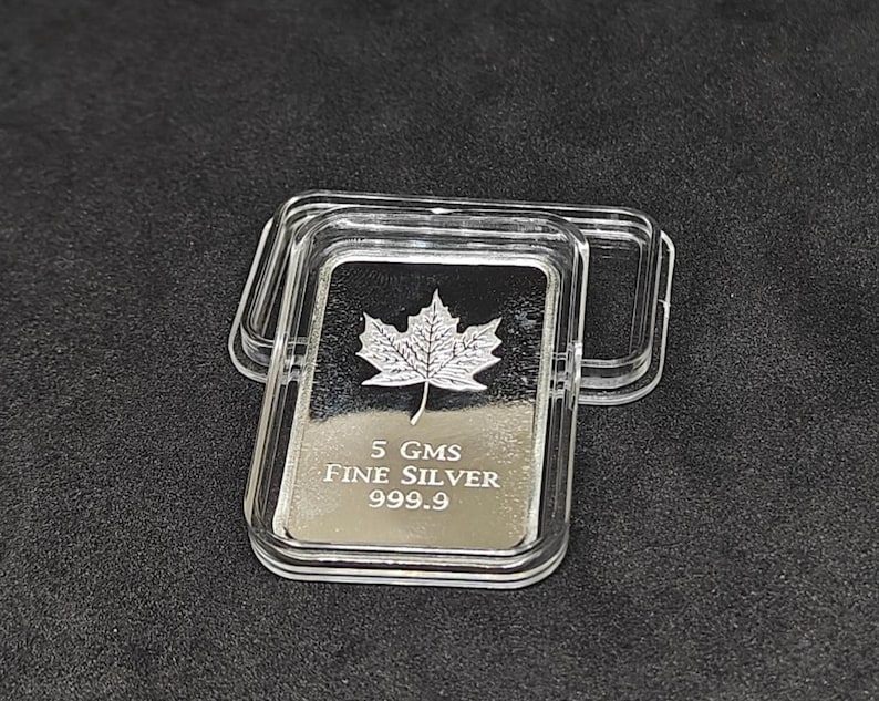 May include: A rectangular silver bar with a maple leaf design, encased in a clear plastic case. The bar is marked with "5 GMS FINE SILVER 999.9". The case is open, revealing the silver bar on a dark surface.