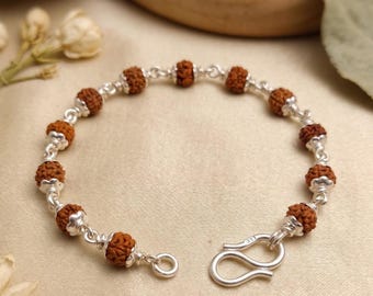 May include: A bracelet featuring eleven small, round, brown, textured rudraksha beads alternating with polished silver links. The piece includes a silver hook-and-eye clasp with a small 925 stamp visible on the metal surface of the hook component.