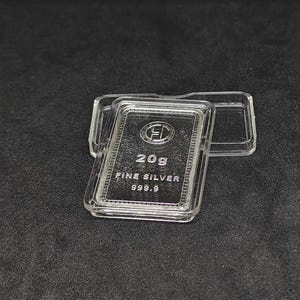 May include: A 20g fine silver bar, marked 999.9, is displayed in a clear protective case. The bar features an embossed logo and text. The case is open, revealing the silver bar against a dark background.