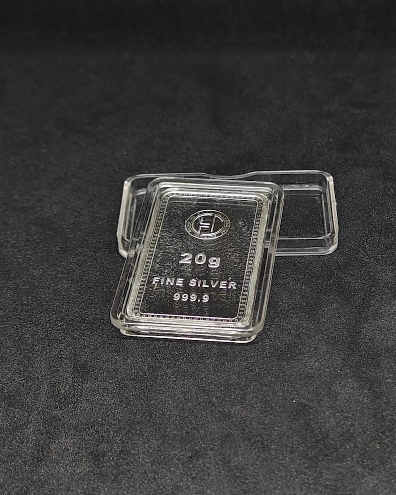 May include: A 20g fine silver bar, marked 999.9, is displayed in a clear protective case. The bar features an embossed logo and text. The case is open, revealing the silver bar against a dark background.