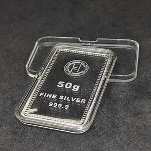 May include: A rectangular silver bar, weighing 50 grams, encased in a clear plastic case. The bar is stamped with the words "FINE SILVER 999.9" and a logo. The case is open, revealing the silver bar against a dark background.