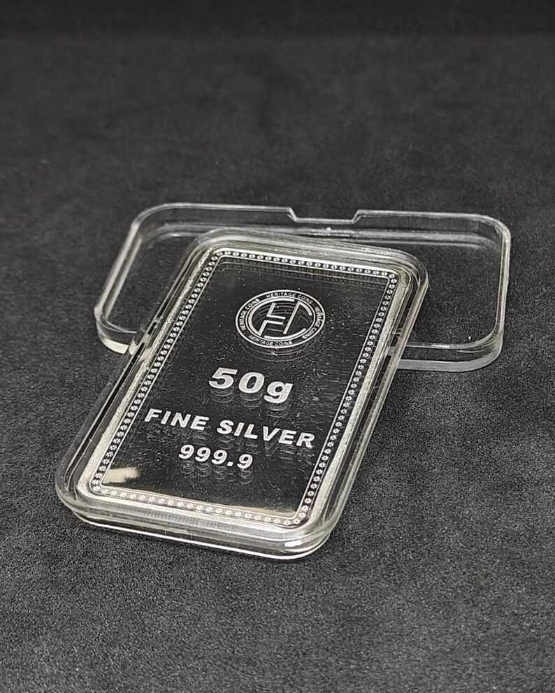 May include: A rectangular silver bar, weighing 50 grams, encased in a clear plastic case. The bar is stamped with the words "FINE SILVER 999.9" and a logo. The case is open, revealing the silver bar against a dark background.
