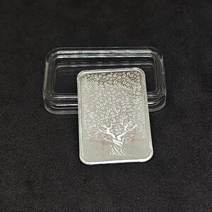 May include: A rectangular silver bar with a tree design, displayed in a clear plastic case. The tree features a detailed trunk and branches with numerous leaves. The bar is on a black surface.
