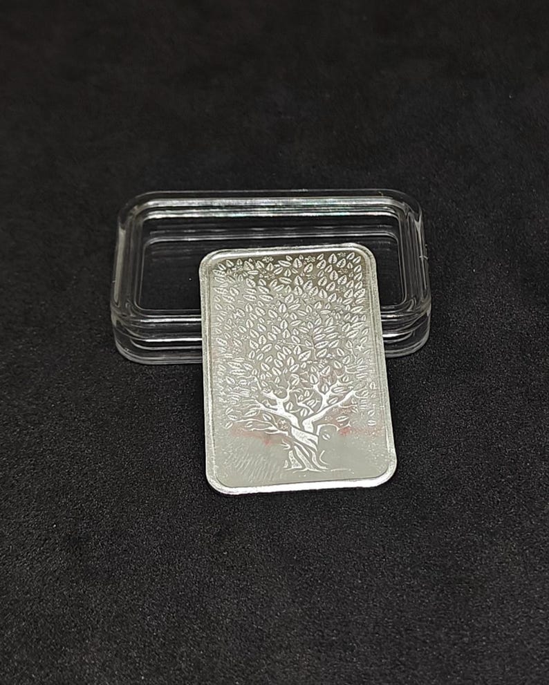 May include: A rectangular silver bar with a tree design, displayed in a clear plastic case. The tree features a detailed trunk and branches with numerous leaves. The bar is on a black surface.