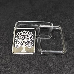 May include: A silver bar with a tree of life design, displayed in a clear plastic case. The tree features a detailed trunk and leafy branches. The bar is rectangular and has a reflective surface, set against a dark background.
