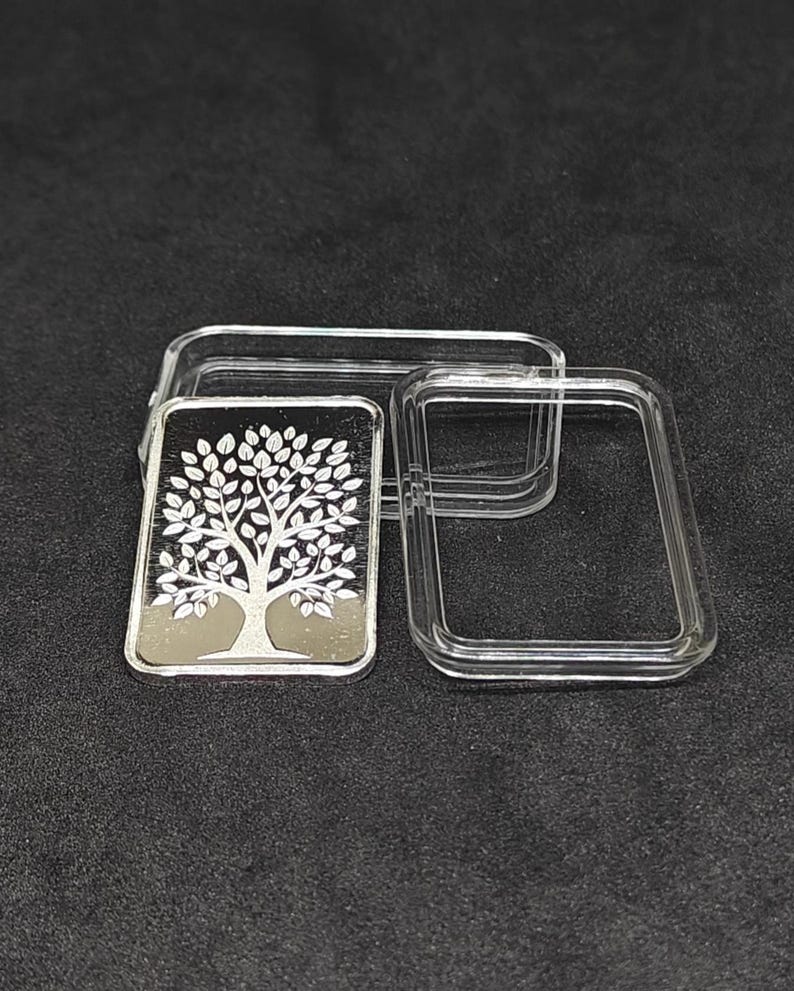 May include: A silver bar with a tree of life design, displayed in a clear plastic case. The tree features a detailed trunk and leafy branches. The bar is rectangular and has a reflective surface, set against a dark background.