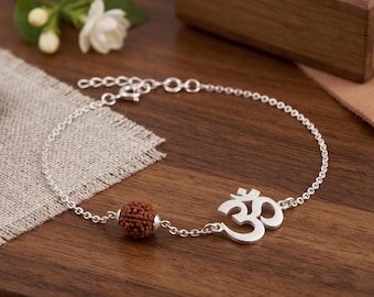 May include: A silver chain bracelet featuring a polished silver Om symbol charm and a single round, textured brown rudraksha bead. The bracelet includes a delicate link chain with a secure clasp mechanism, showcasing a balanced design.