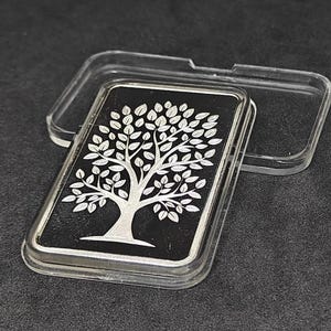 May include: A rectangular silver bar with a tree design on a black background, encased in a clear plastic case. The tree is depicted with intricate details, including branches and leaves. The bar is presented on a dark surface.