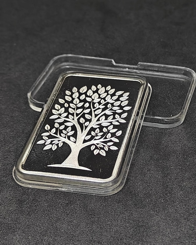 May include: A rectangular silver bar with a tree design on a black background, encased in a clear plastic case. The tree is depicted with intricate details, including branches and leaves. The bar is presented on a dark surface.