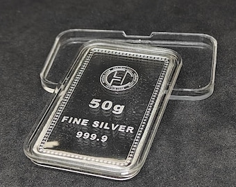 May include: A rectangular silver bar, marked "50g FINE SILVER 999.9", is displayed in a clear plastic case. The bar has a detailed border and a logo. The case is open, revealing the silver bar against a dark background.