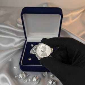 May include: A silver coin featuring a profile of Queen Victoria is held in a black gloved hand. The coin is displayed against a backdrop of a blue velvet box and scattered diamonds on a satin surface. The coin is labeled "VICTORIA QUEEN".