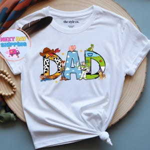 May include: White t-shirt with the word "DAD" in colorful, cartoon-style letters. The letters are designed with characters from the Toy Story movie. The shirt has a crew neck and short sleeves.