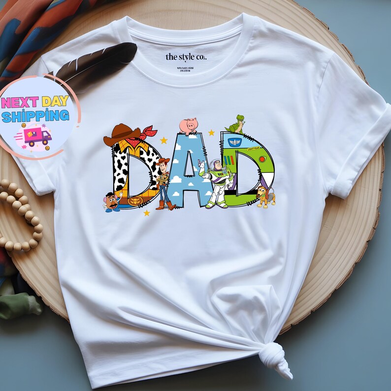 May include: White t-shirt with the word "DAD" in colorful, cartoon-style letters. The letters are designed with characters from the Toy Story movie. The shirt has a crew neck and short sleeves.