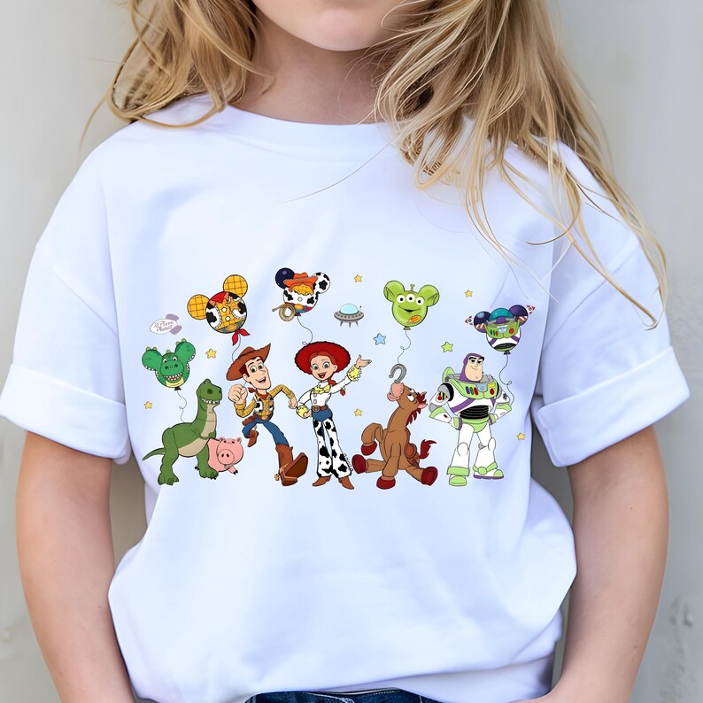 May include: White t-shirt with a vibrant graphic of Toy Story characters. The design includes Woody, Buzz Lightyear, Jessie, and other characters, along with balloons and stars. The shirt is made of a soft material.